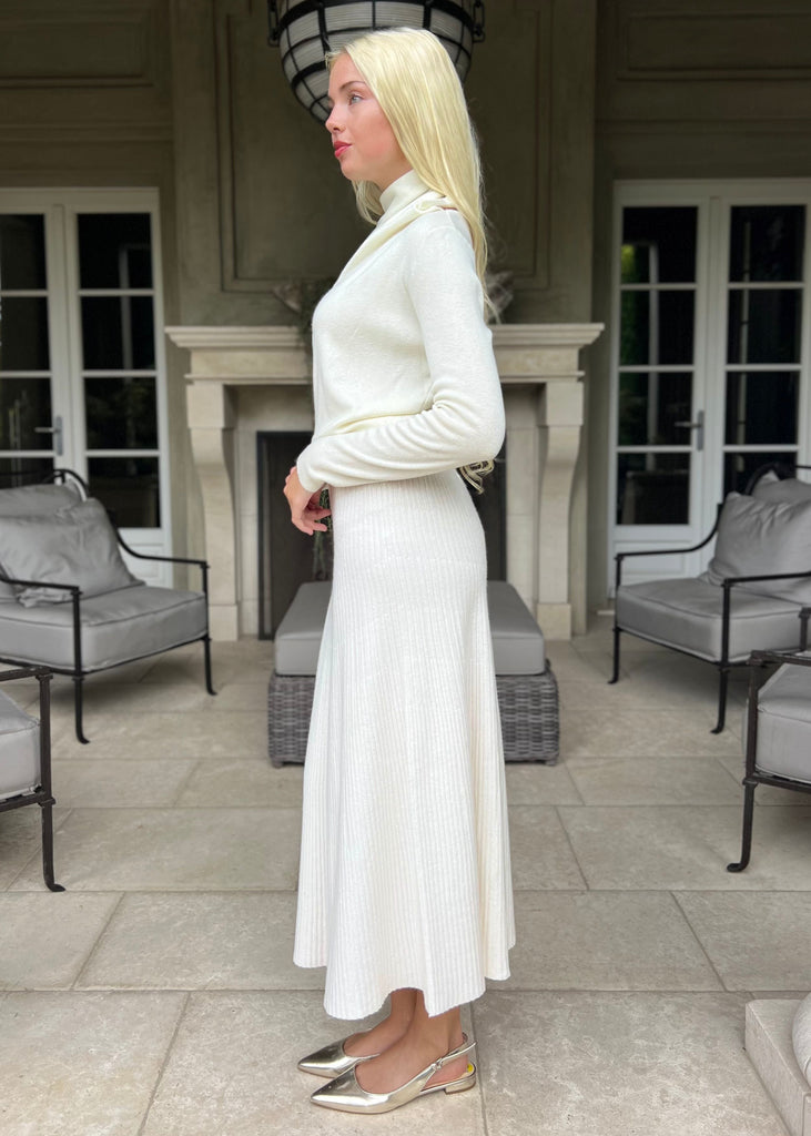 Woman in a white outfit standing in a stylish outdoor setting with chairs and a fireplace.