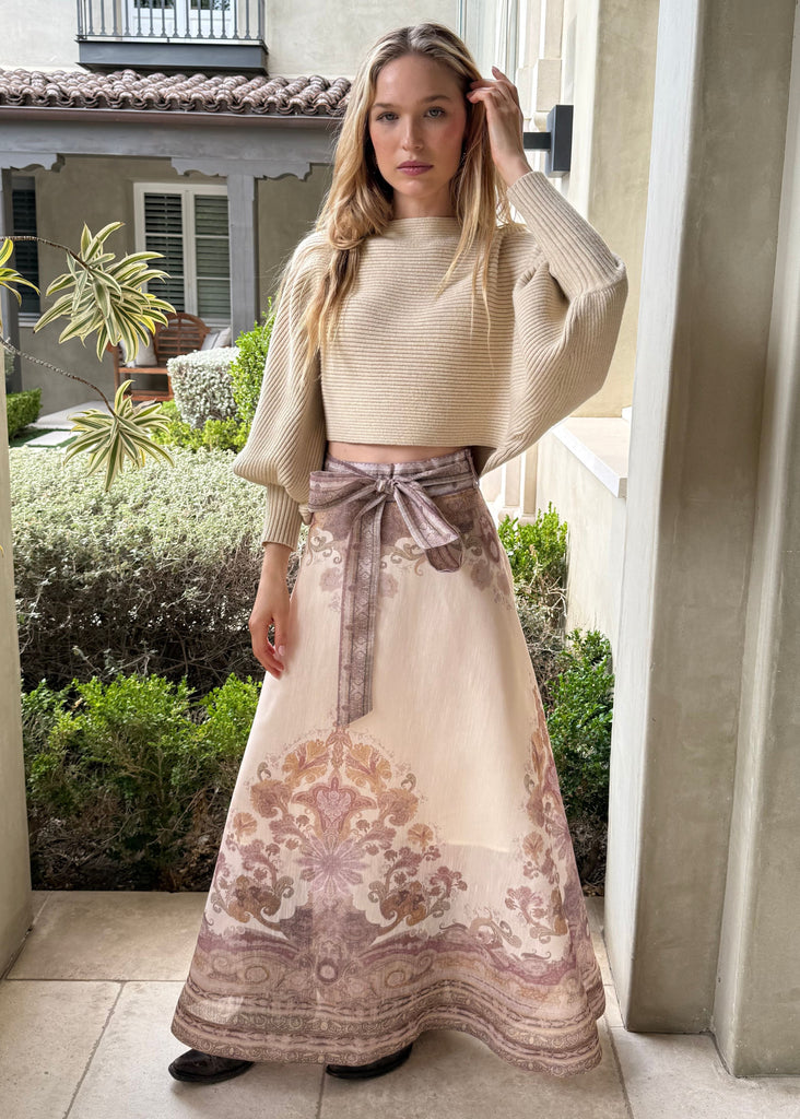 Woman in a beige sweater and patterned skirt standing in a doorway.
