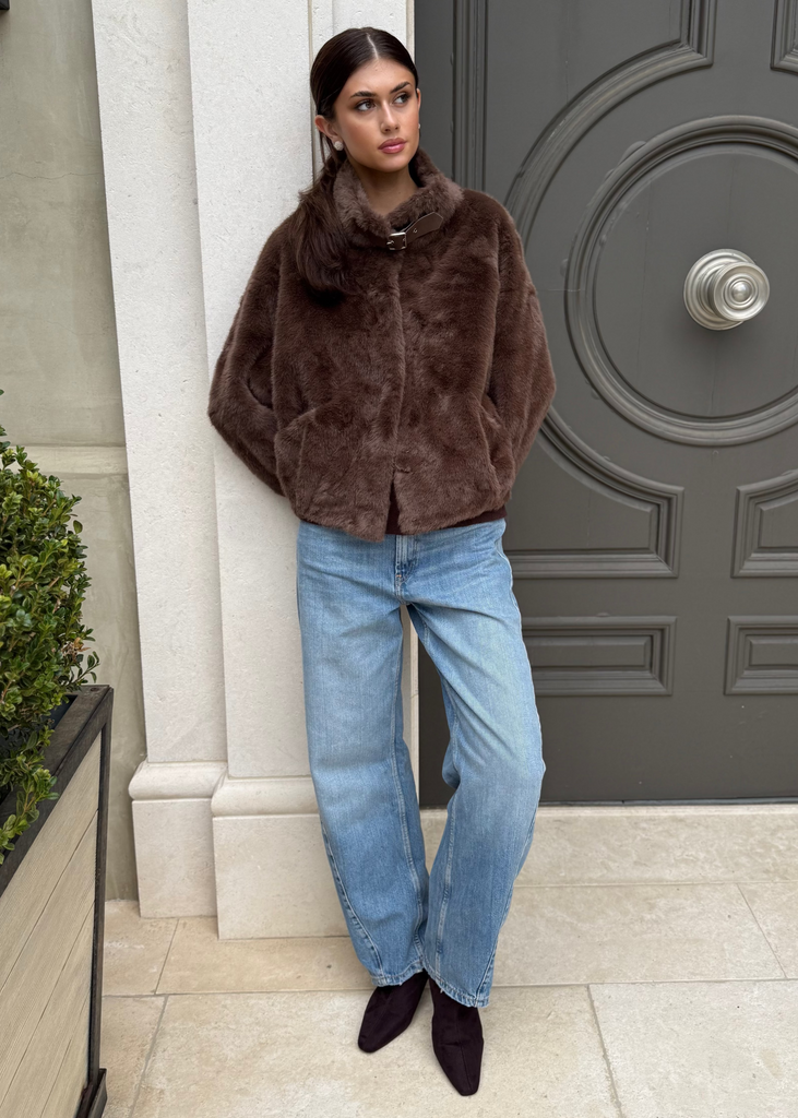 Woman in a brown fur coat and blue jeans standing in front of a large door.