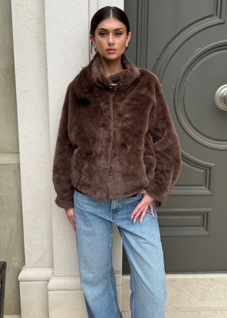 Woman wearing a brown fur coat and blue jeans standing in front of a gray door.