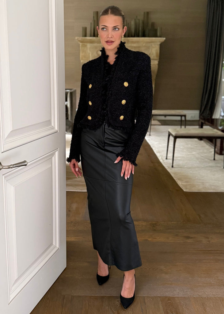 Woman in a black coat with gold buttons and black skirt standing in a doorway.