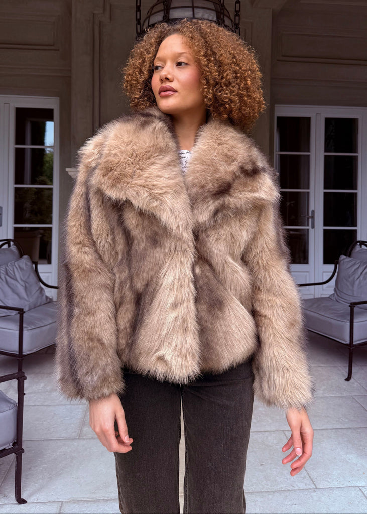 Woman wearing a fur coat in an elegant indoor setting with white chairs and a chandelier.