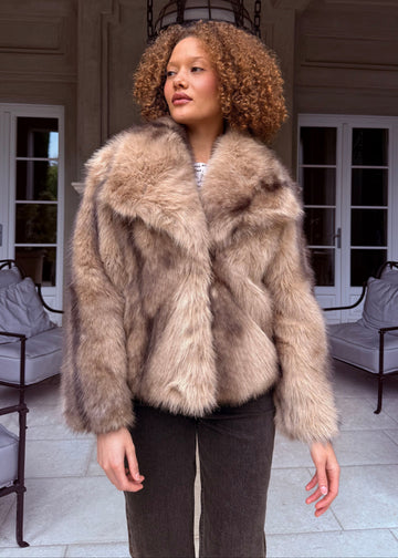 Woman wearing a fur coat in an elegant indoor setting with white chairs and a chandelier.