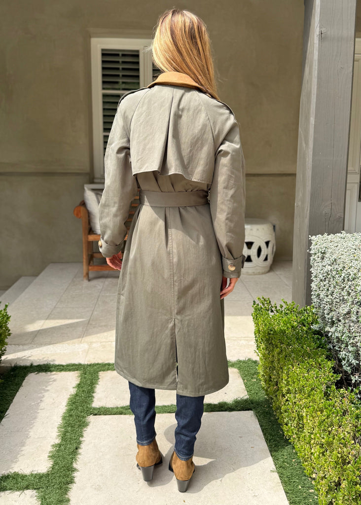 Person wearing a long coat walking on a patio with greenery and a building in the background