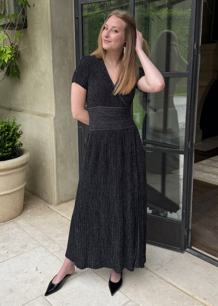Woman in a black dress standing outdoors next to a building with plants.