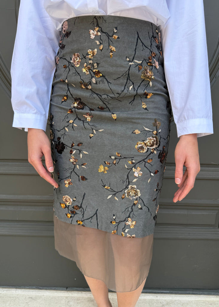 Person wearing a gray skirt with floral embroidery in front of a dark green door.
