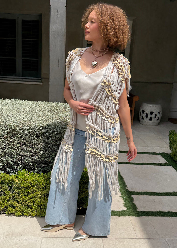 Woman standing outdoors in a garden setting wearing a white top and light blue jeans with a decorative beige cardigan.