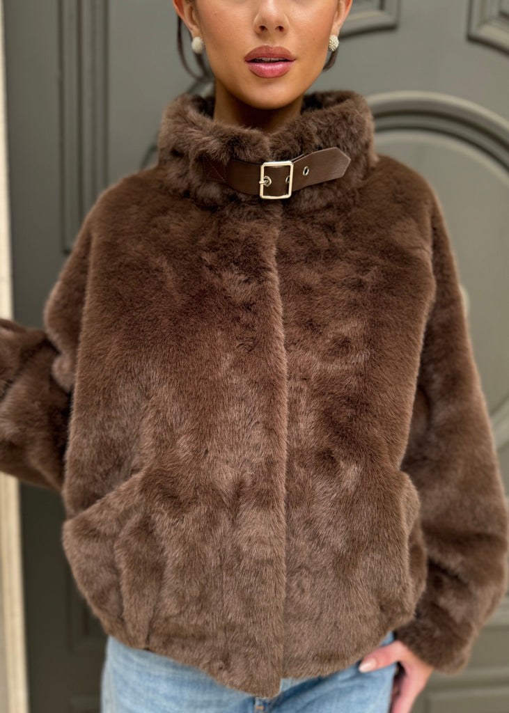 Woman wearing a brown fur jacket with a belt, standing against a neutral background.