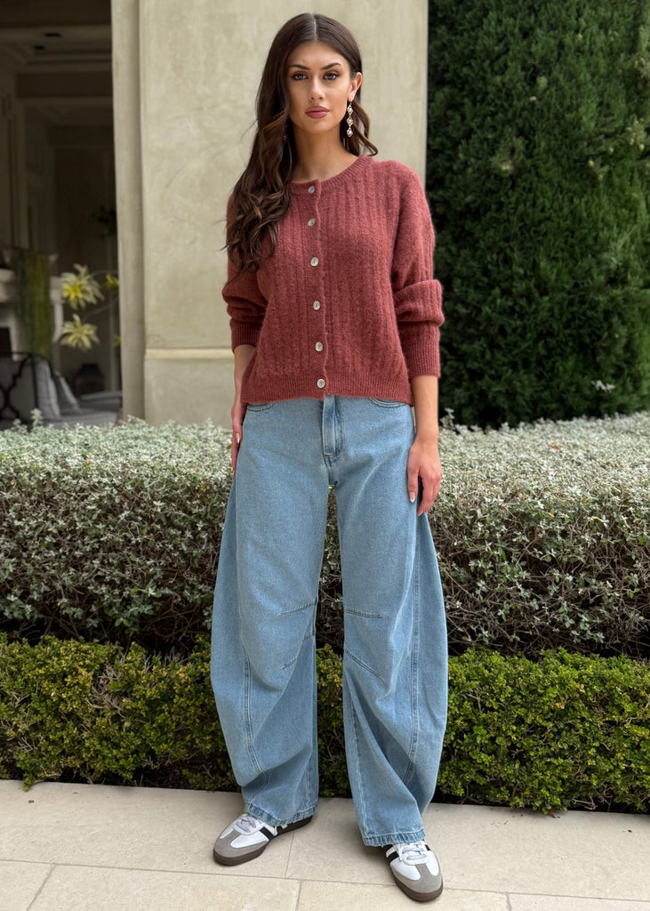 Woman in a red sweater and blue jeans standing in front of a building with greenery.