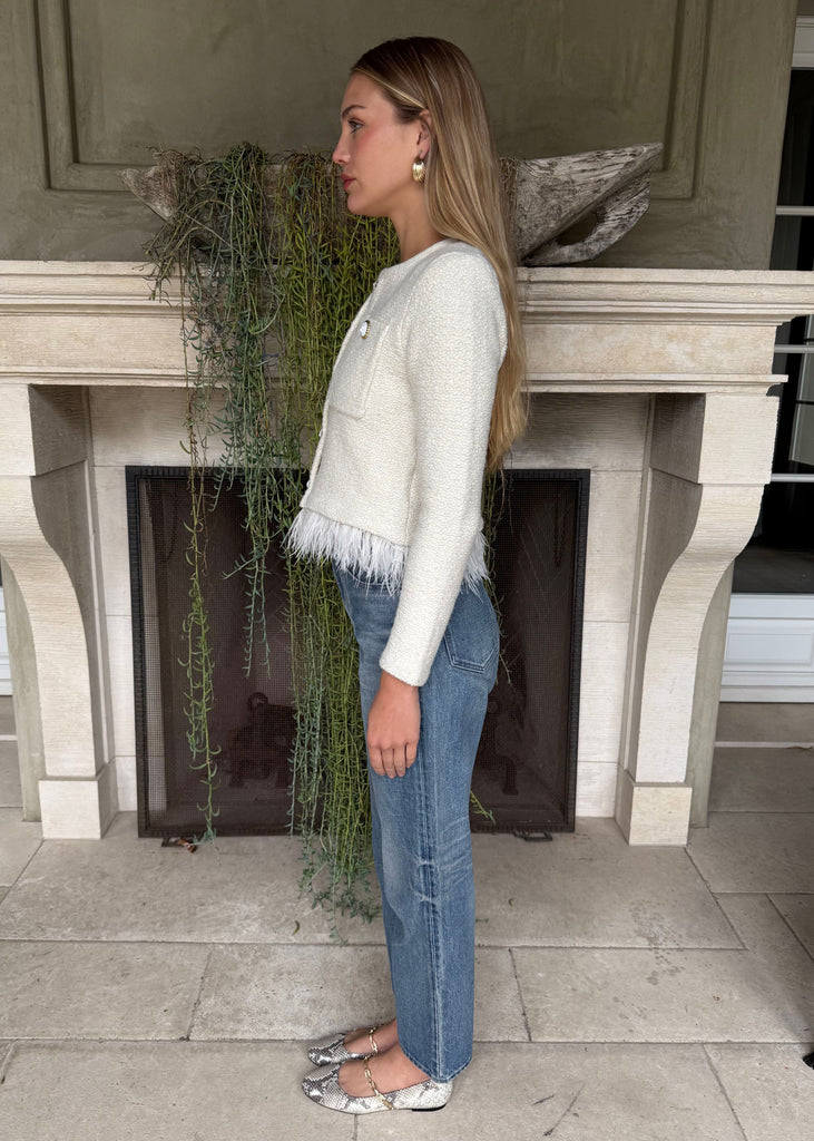 Woman standing in front of a fireplace with a plant, wearing a white sweater and blue jeans.
