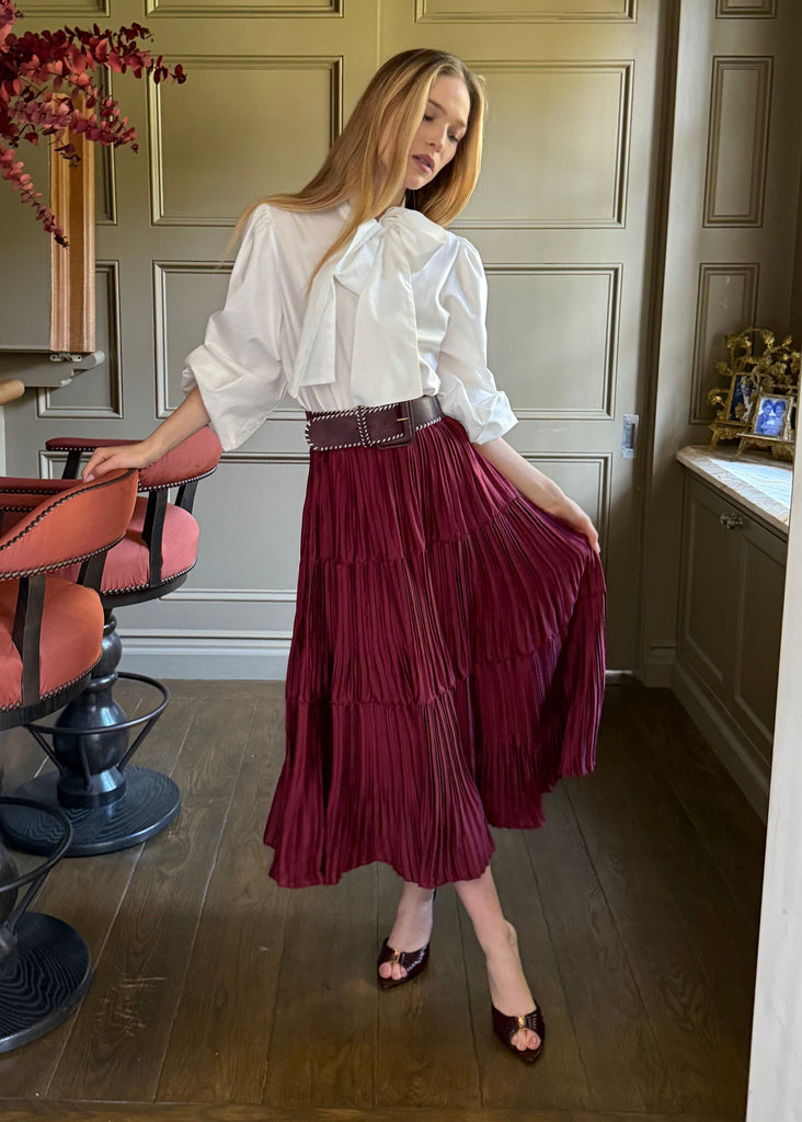Woman in a white blouse and burgundy skirt standing in a room with wooden paneling and a table with flowers.