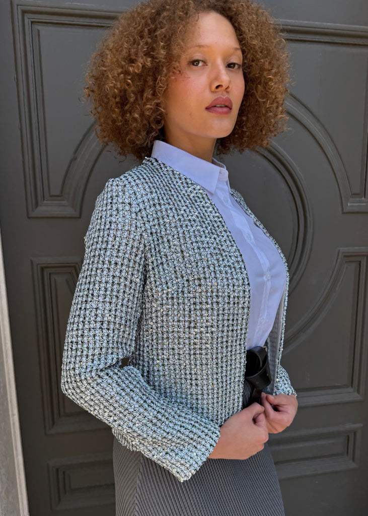 Woman wearing a textured gray jacket over a light blue shirt and dark skirt, standing in front of a gray door.