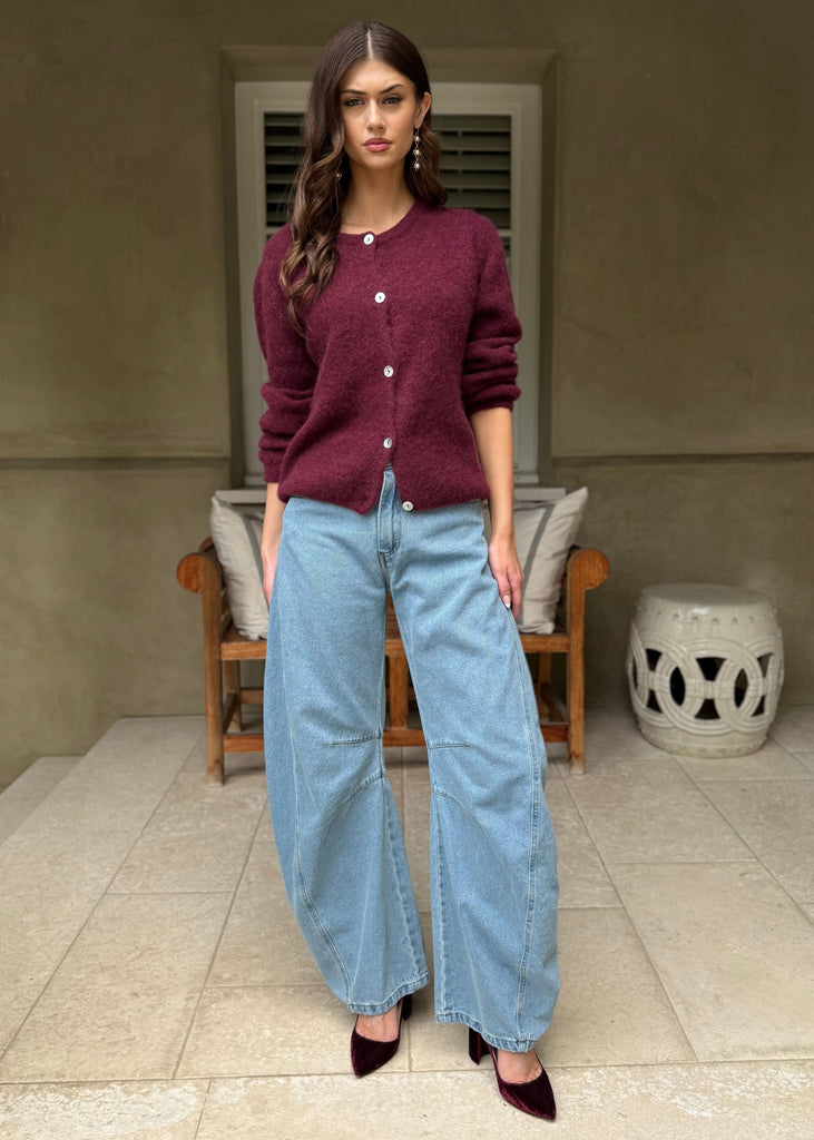 Woman wearing a maroon cardigan and light blue jeans standing in front of a house.