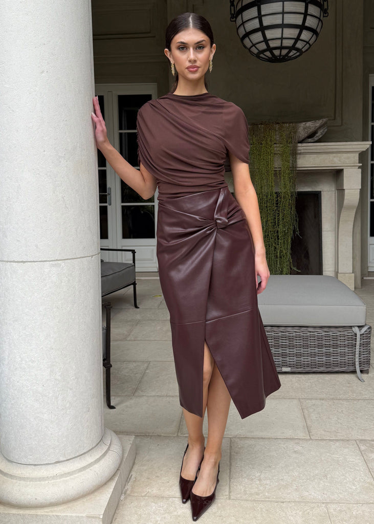 Woman in a brown dress standing in a stylish outdoor setting with columns and furniture.