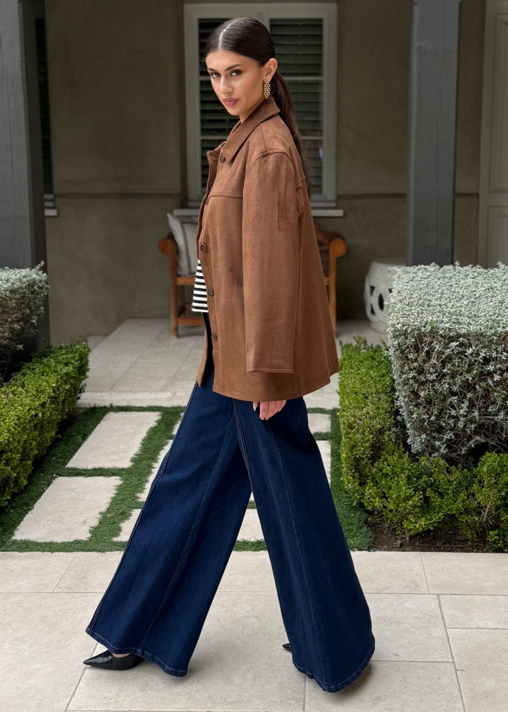 Woman in a brown jacket and blue pants walking in front of a house with a garden.