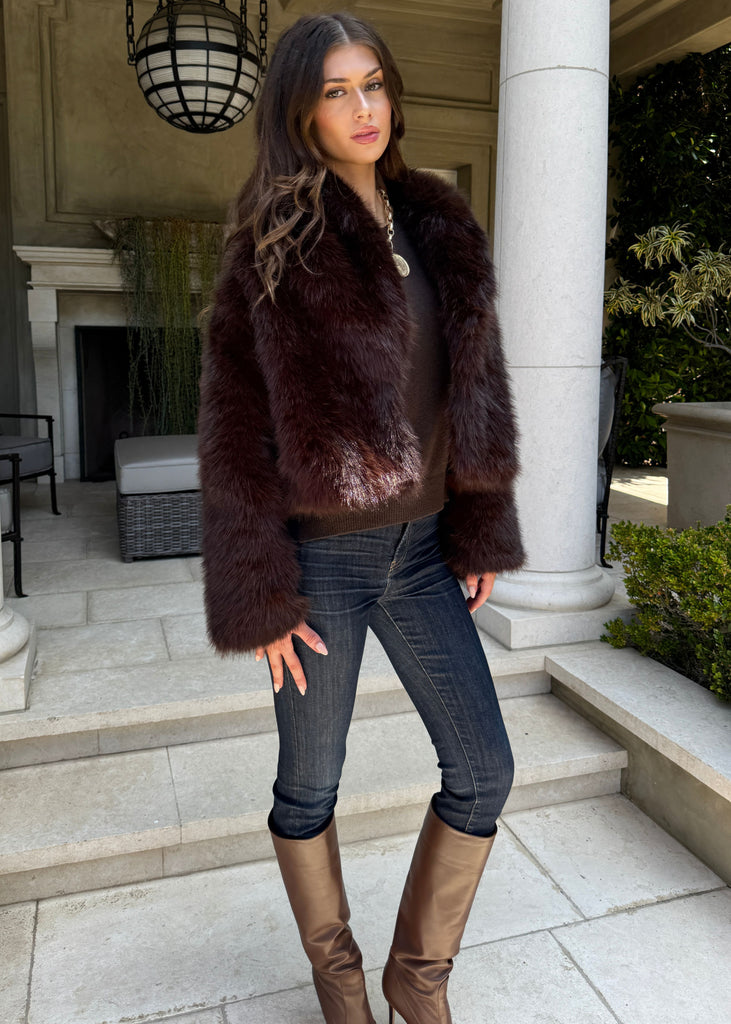 Woman wearing a brown fur coat, dark jeans, and brown boots standing in front of a building with columns.
