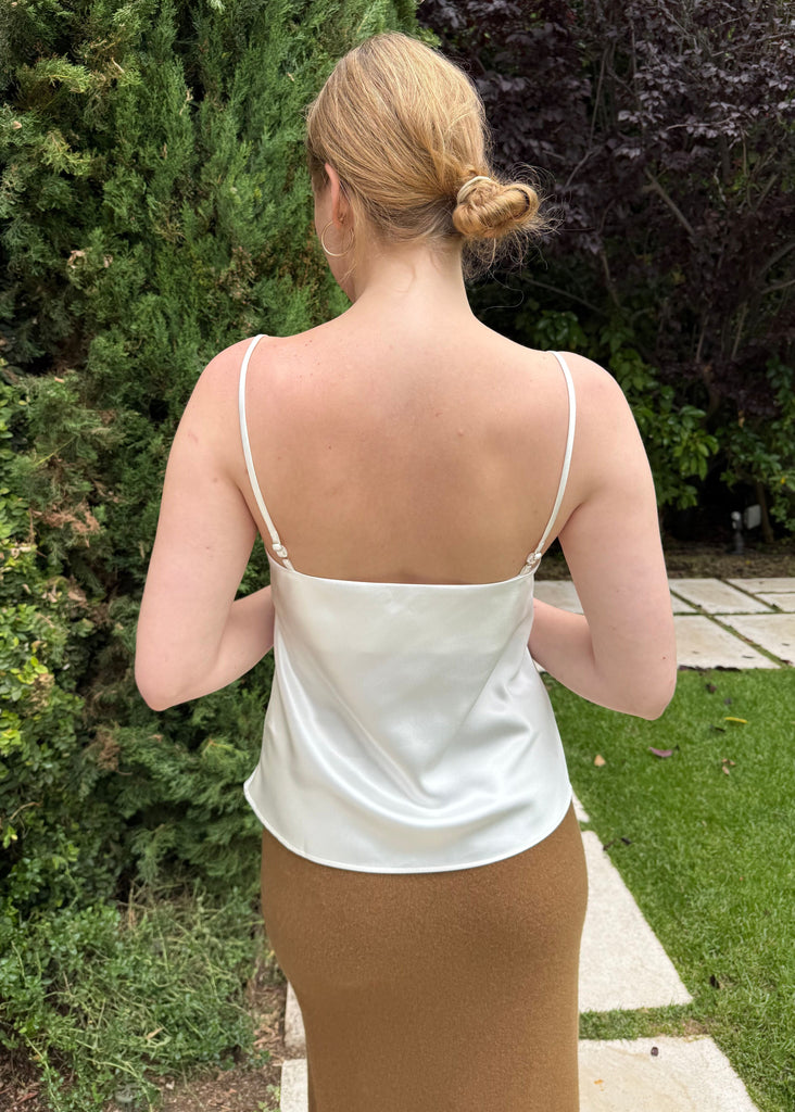 Person wearing a white top and brown skirt standing outdoors with greenery in the background