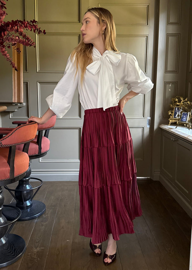 Woman in a white blouse and red skirt standing in a room with wooden floors and a decorative plant.