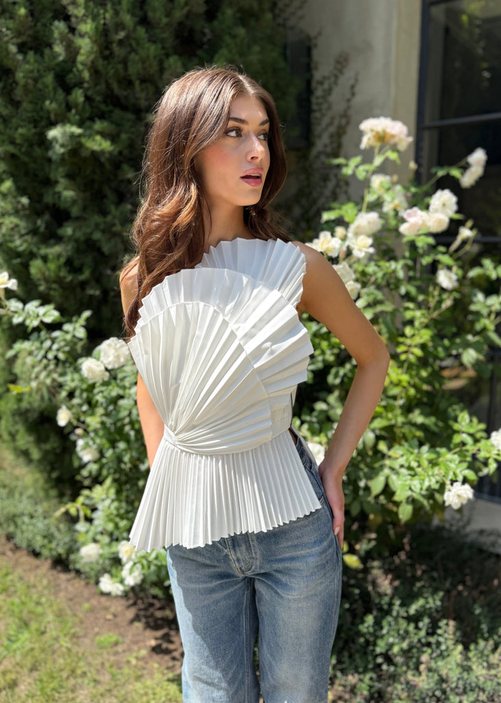 Woman wearing a white pleated top and blue jeans standing in a garden with greenery and flowers.