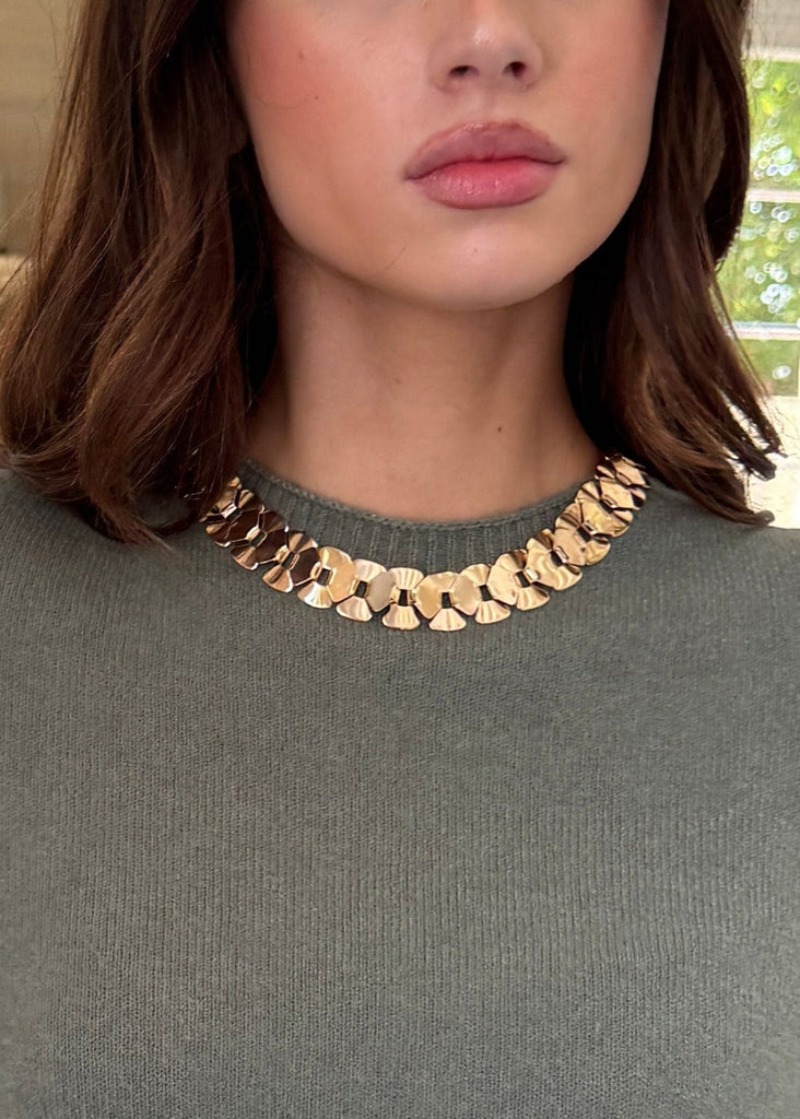 Woman wearing a green sweater and gold necklace in a room with a fireplace.