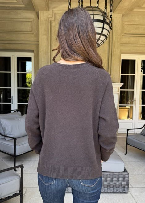 Person wearing a gray sweater and blue jeans standing on a patio with chairs and a lamp in the background.