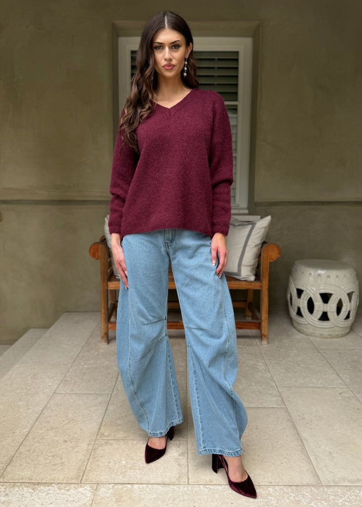 Woman wearing a burgundy sweater and light blue jeans standing in a room with a neutral color scheme.