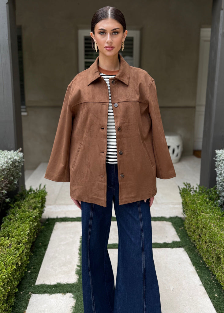 Woman wearing a brown jacket, striped shirt, and blue jeans standing in a garden.