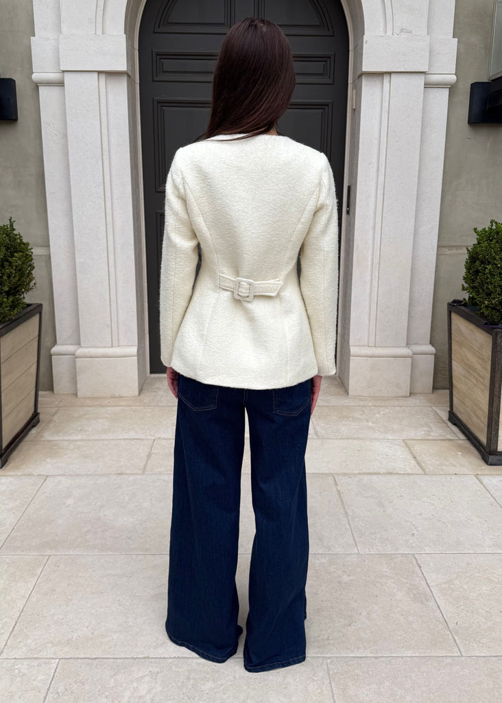Person wearing a white blazer and blue pants standing in front of a building entrance.