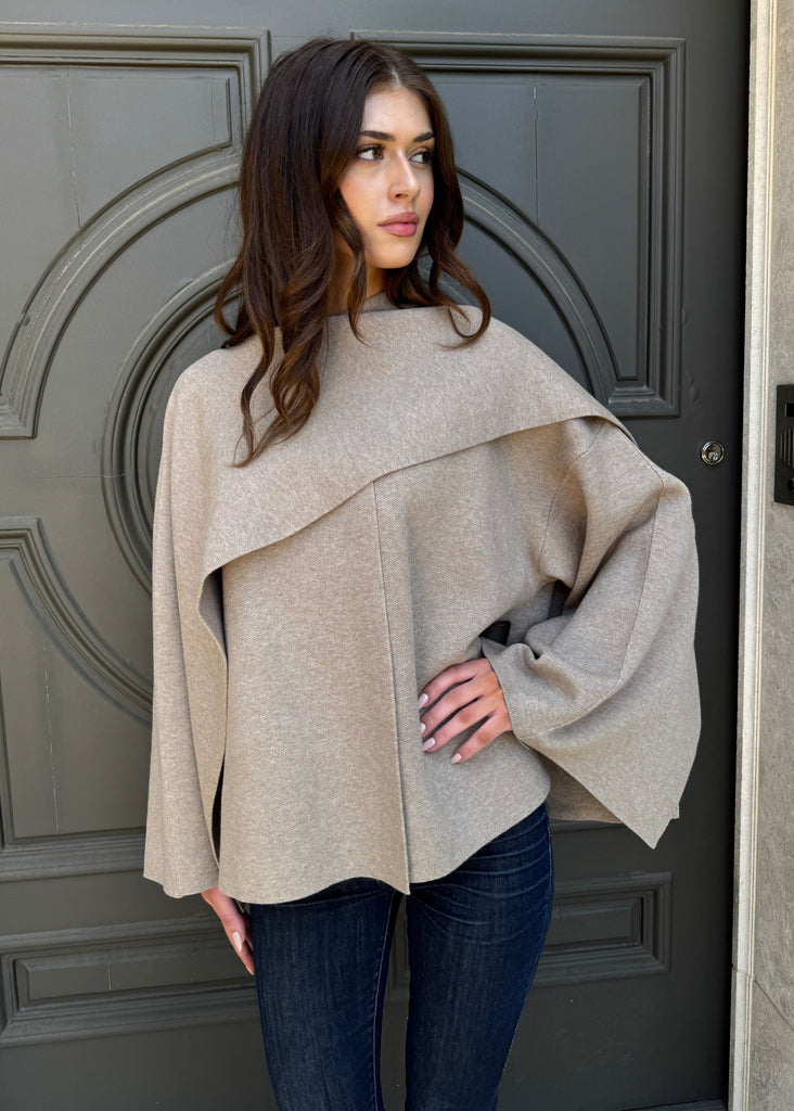 Woman wearing a beige poncho standing in front of a gray door.