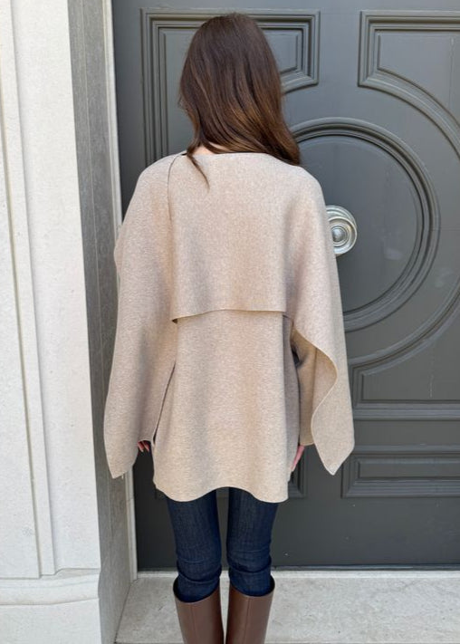 Person wearing a beige poncho standing in front of a gray door.