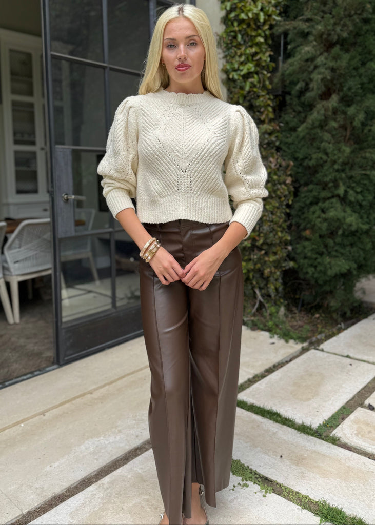 Woman wearing a cream sweater and brown leather pants standing outdoors.