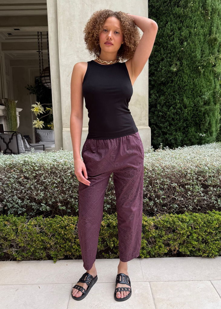 Woman wearing a black sleeveless top and purple pants standing outdoors.
