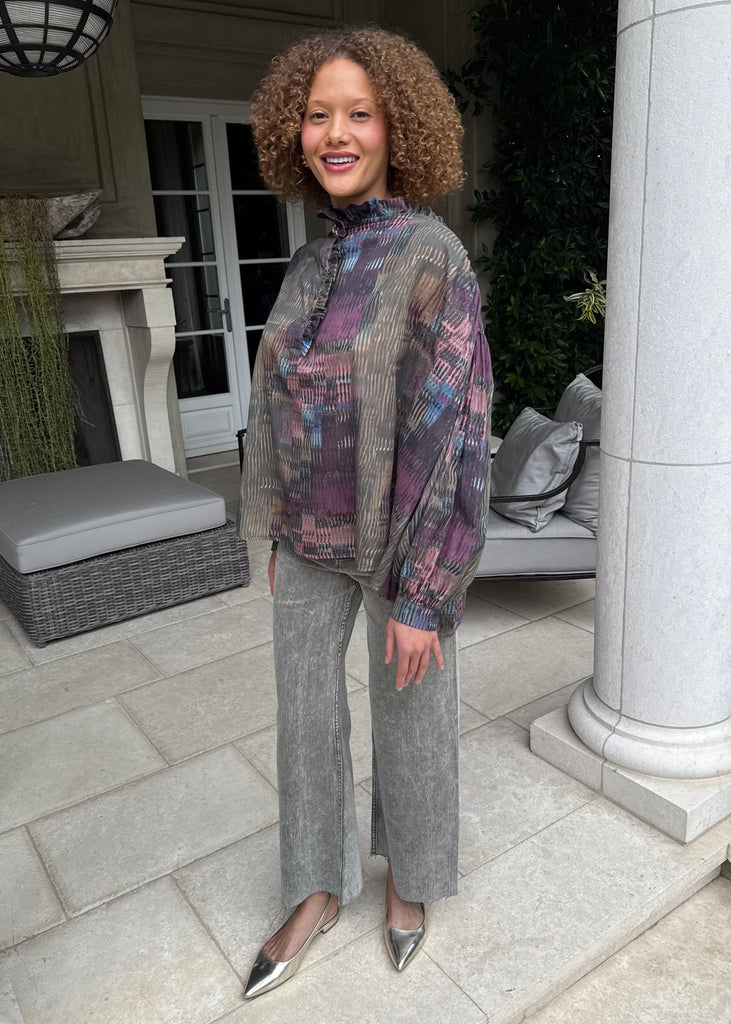 Woman wearing a patterned blouse and gray pants standing outdoors.
