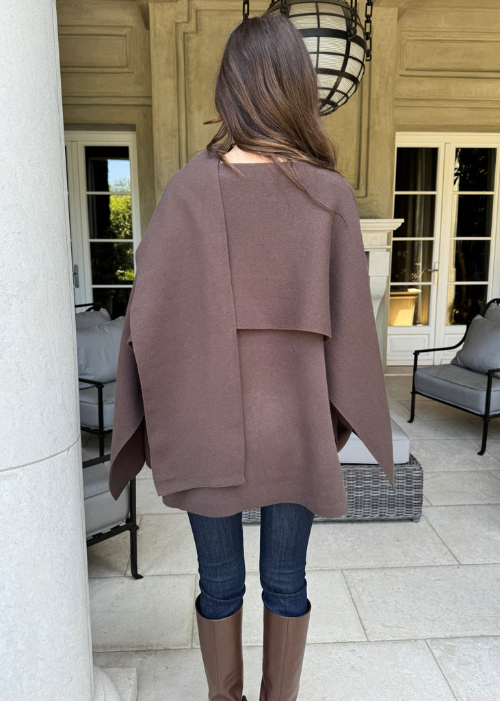 Person wearing a brown cape in an outdoor setting with columns and furniture.