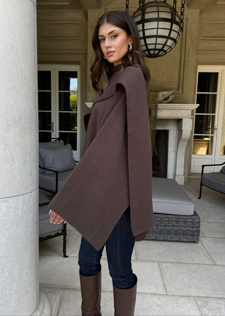 Woman wearing a brown cape in an outdoor setting with columns and furniture.