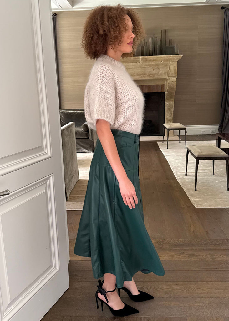 Woman in a green skirt and white top standing in a well-lit room with wooden flooring.
