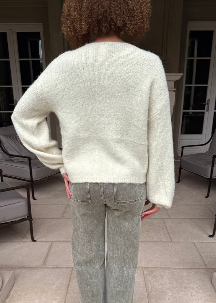 Person wearing a light-colored sweater and gray pants standing in an outdoor setting with columns and chairs.