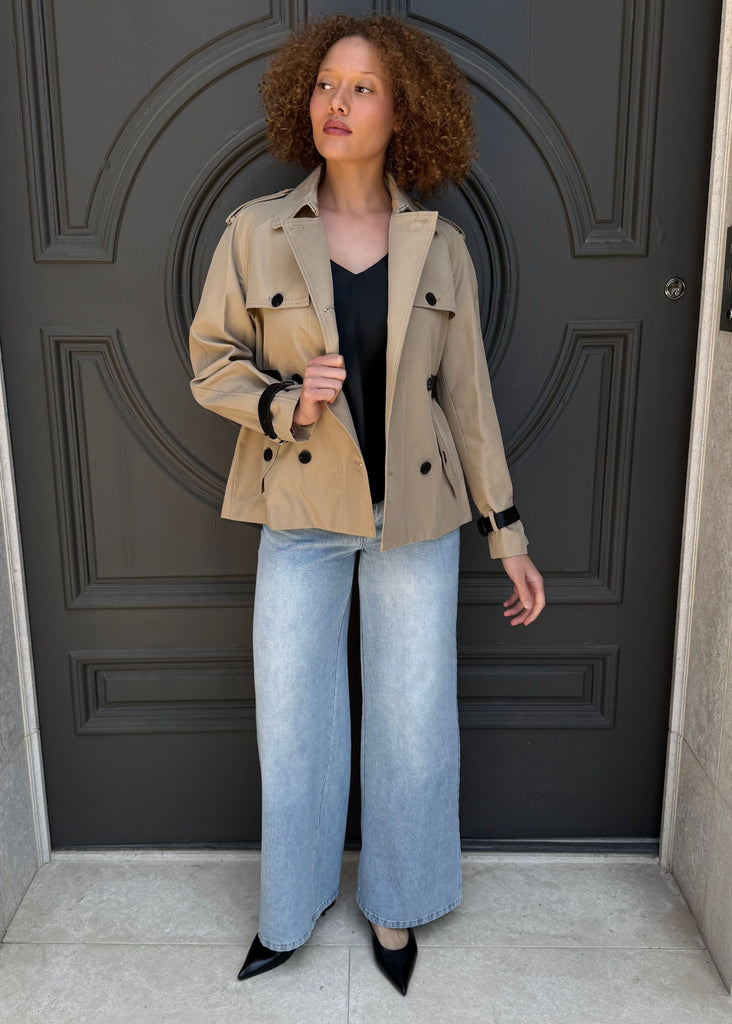 Woman standing in front of a dark gray door wearing a beige jacket, black top, and light blue jeans.