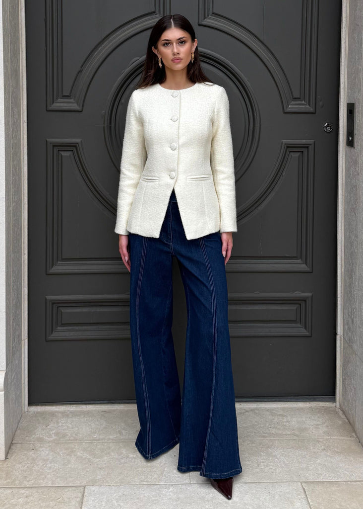 Person wearing a white blazer and blue pants standing in front of a large gray door.