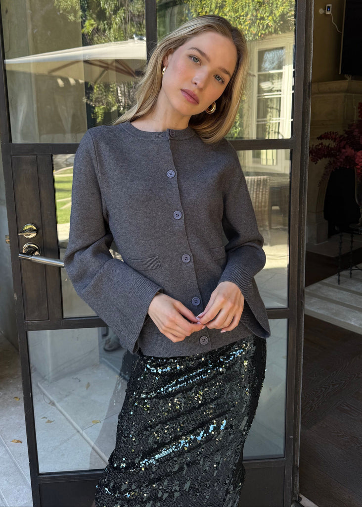 Woman standing in a doorway wearing a gray jacket and black sequin skirt.