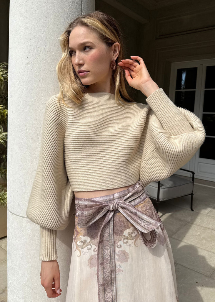Woman in a beige sweater and patterned skirt standing against a building exterior.
