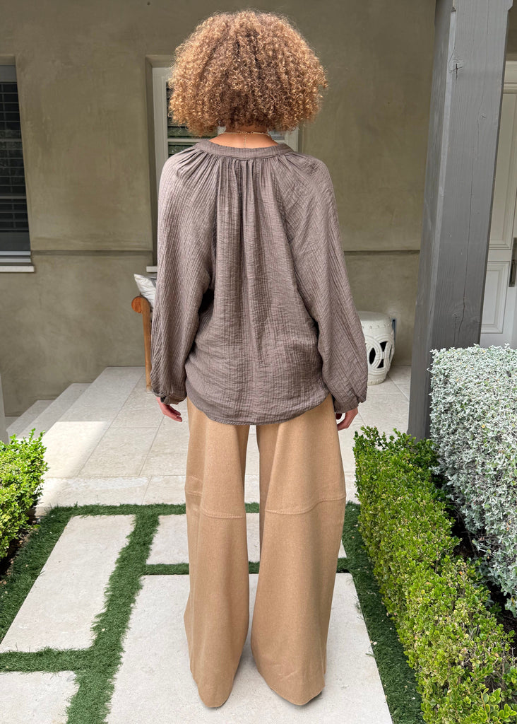 Person wearing a brown top and beige pants standing in a garden with greenery and a building in the background.