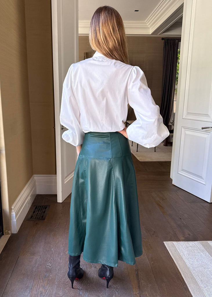 Person wearing a white blouse and teal skirt standing in a room with large windows.