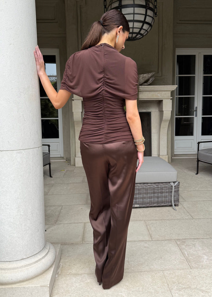 Woman in brown outfit standing in a stylish outdoor setting with columns and furniture.