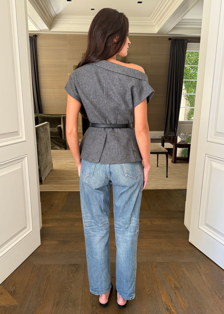 Woman wearing a gray top and blue jeans standing in a room with wooden flooring and white walls.