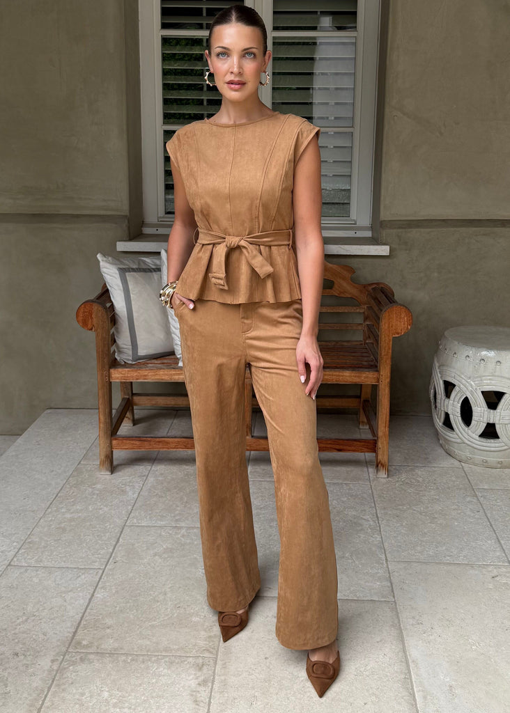 Woman in a brown jumpsuit standing on a patio with a neutral wall and chair in the background.