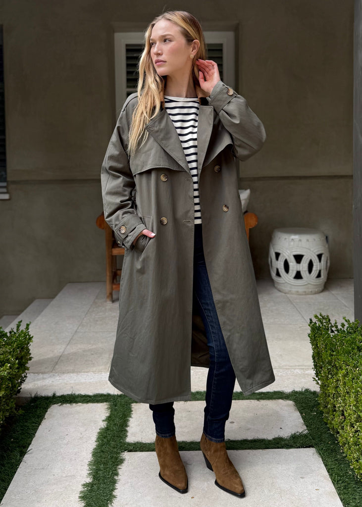 Woman in a gray trench coat standing in a garden setting