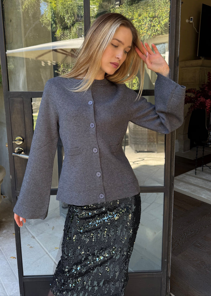Woman wearing a gray cardigan and black sequin skirt standing in front of a glass door.