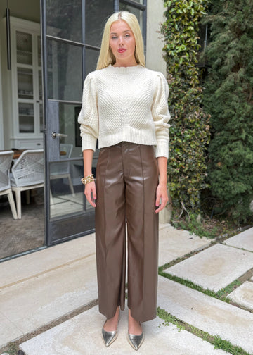 Woman in a white sweater and brown leather pants standing outdoors near a building with large windows.