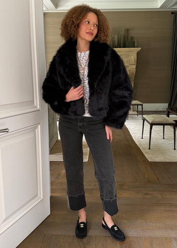 Woman standing in a room wearing a black fur coat, patterned top, and jeans.